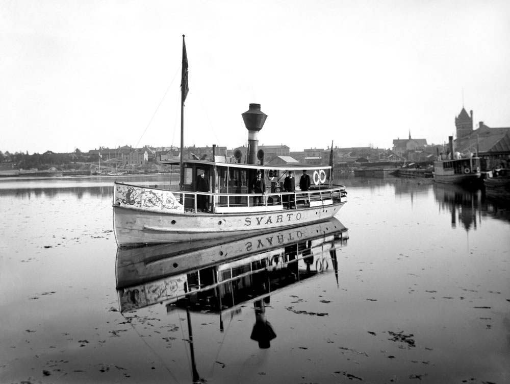 Liten passagerarbåt med skorsten på lugnt vatten framför stad.