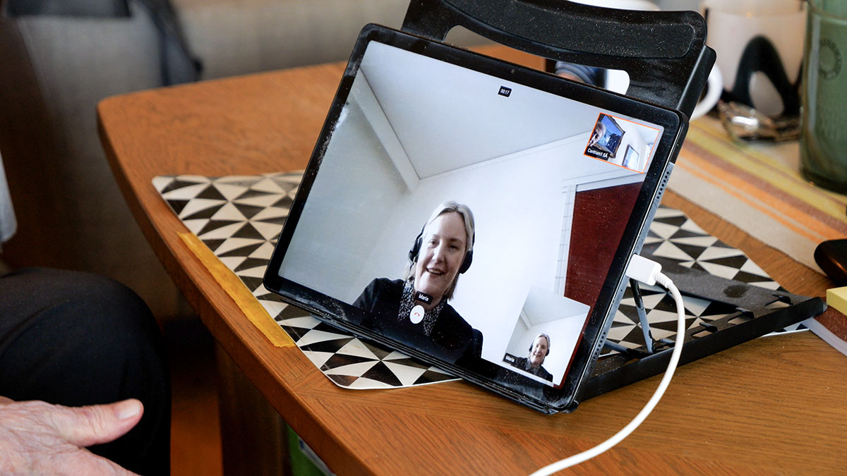 En surfplatta står lutad på ett bord och visar ett videosamtal med en person i hemmiljö. Surfplattan är ansluten med laddkabel och står på en mönstrad duk. I förgrunden syns en hand nära bordskanten.